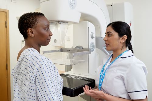 Photo showing a mammographer speaking to a patient, wearing a hospital gown, before a mammogram